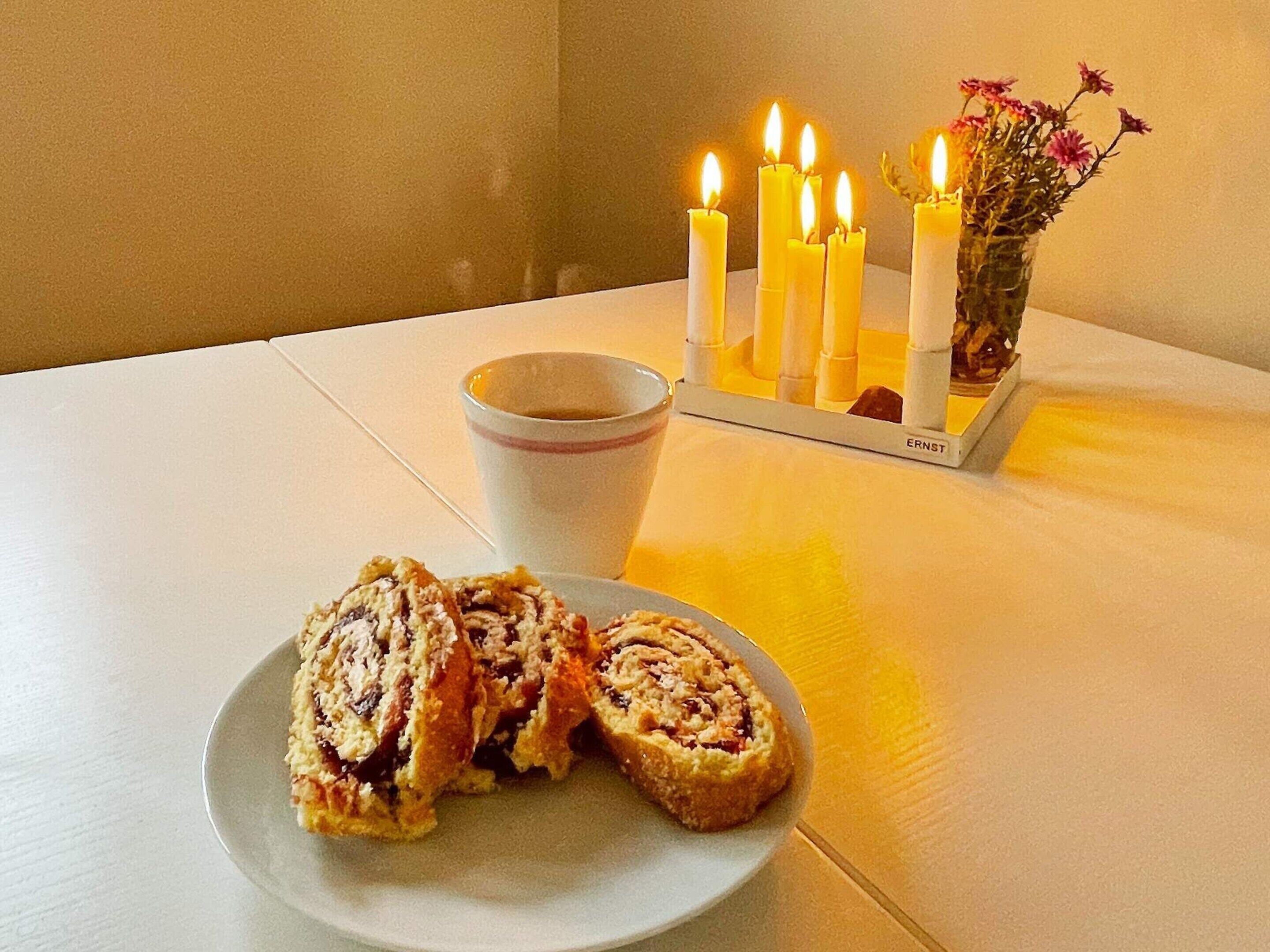 A table with a plate of sliced pastries, a cup of coffee, and a candle holder with lit candles and flowers.