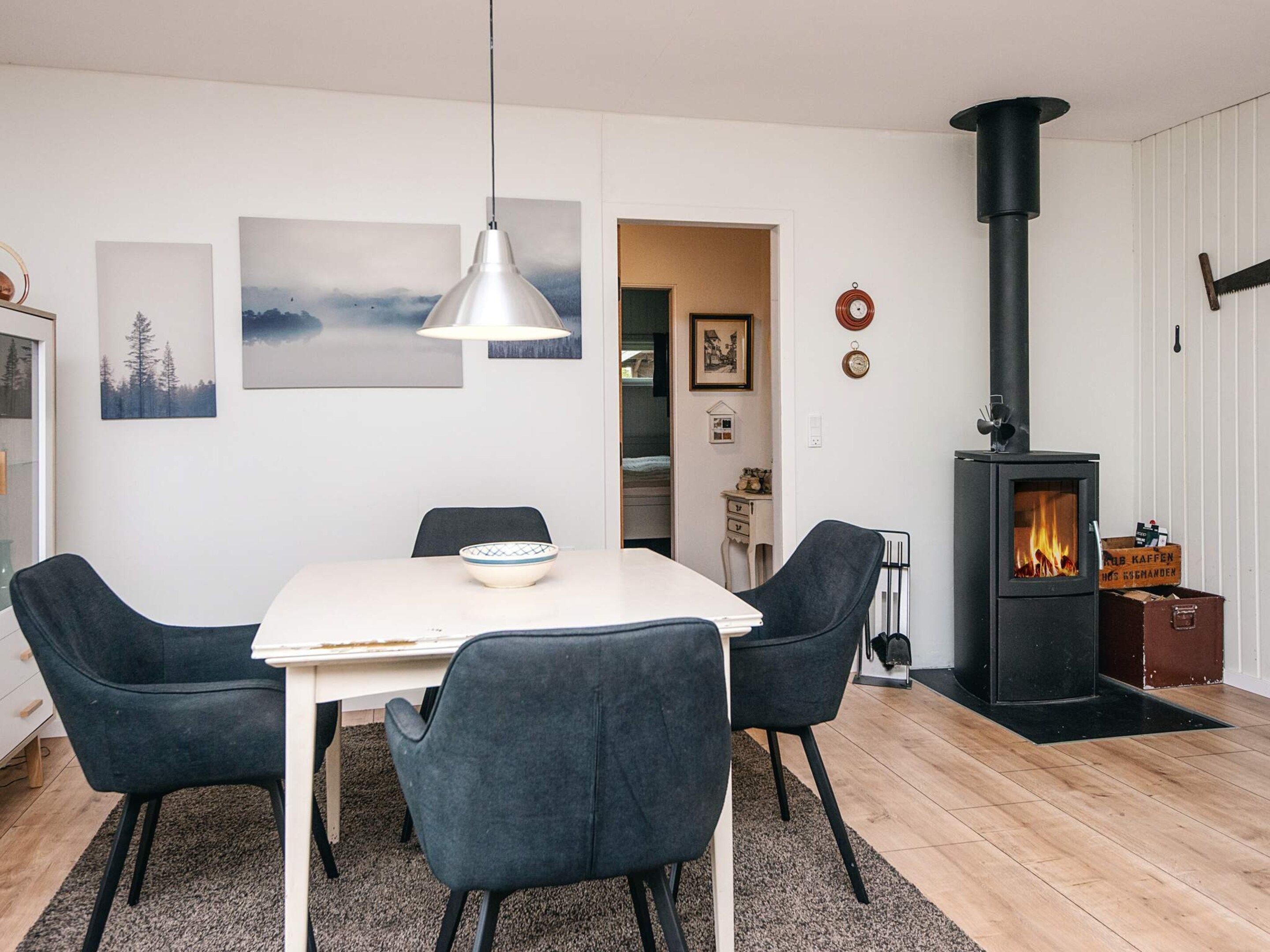Un salon moderne avec une table blanche, quatre chaises foncées, un luminaire suspendu, une cheminée et des tableaux encadrés accrochés au mur.