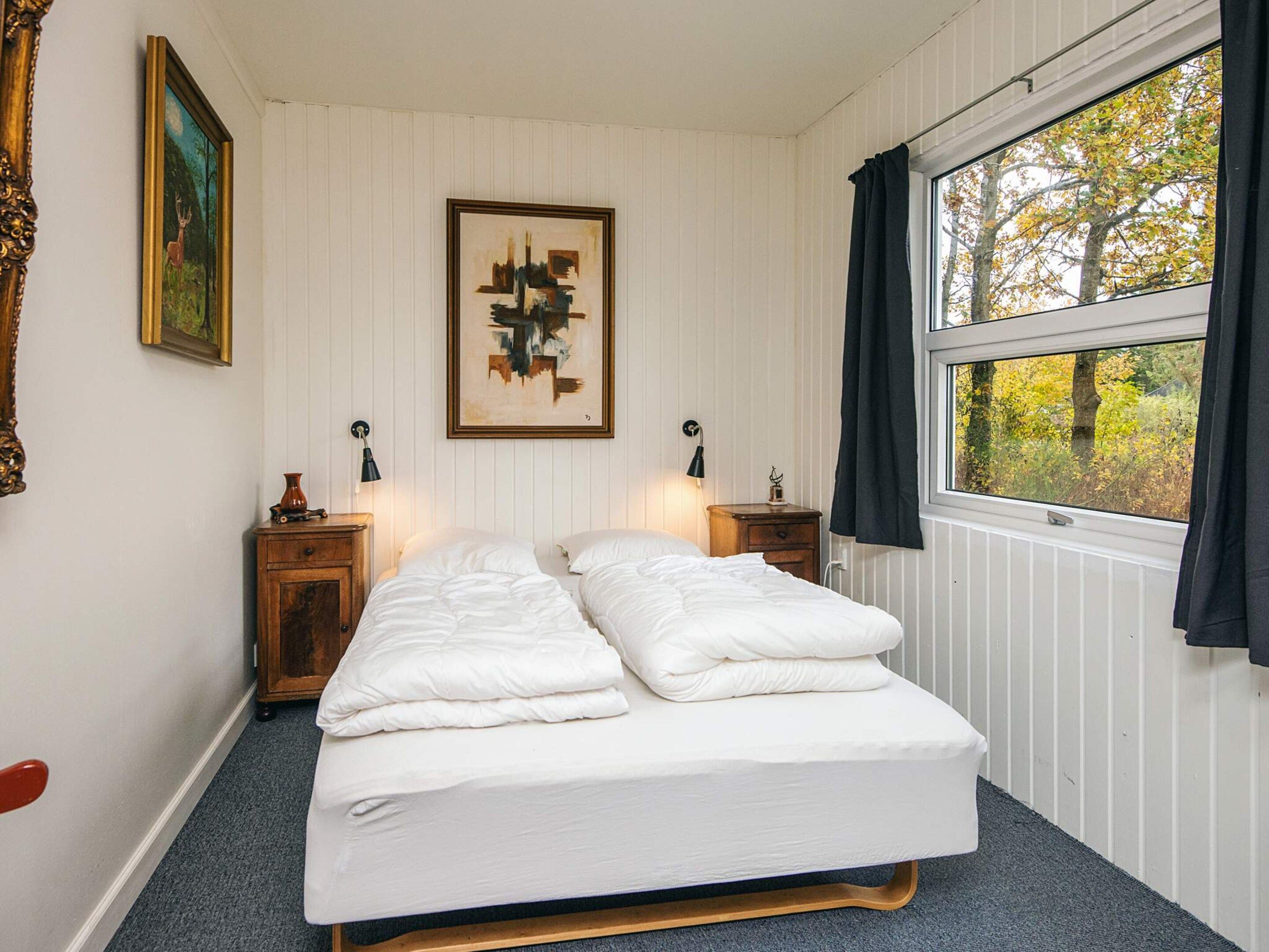 Un dormitorio con cama, dos mesitas de noche, una lámpara de pared, un cuadro abstracto enmarcado y una ventana con cortinas.