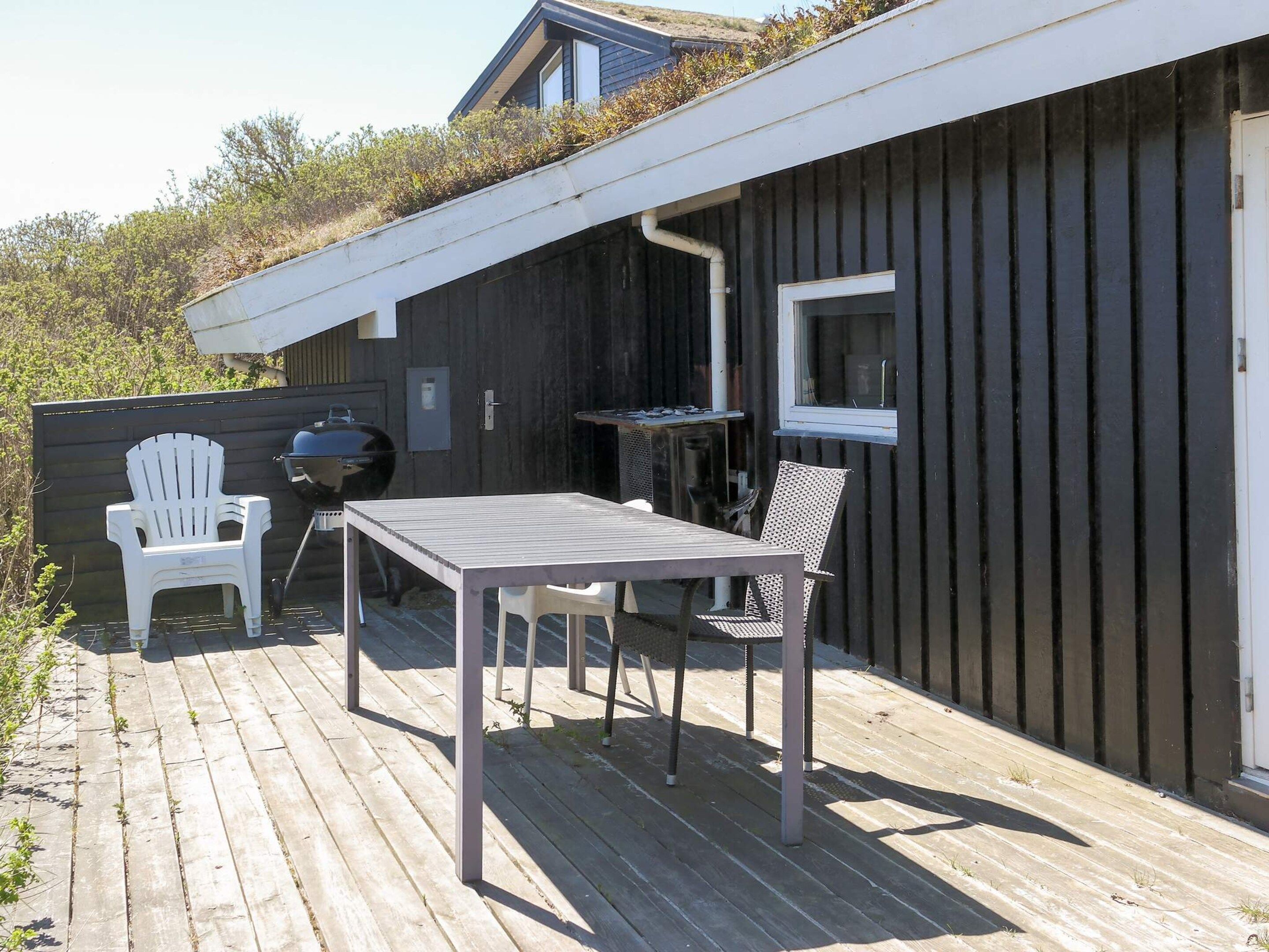 A wooden deck with a table and chairs, a grill, and a window overlooking a garden.