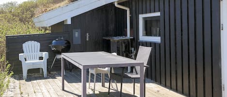 Una terraza de madera con una mesa, sillas, una parrilla y una ventana que da a un jardín.