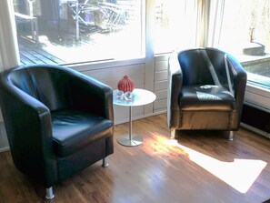 A room with two black armchairs, a small round table with a red vase, and a view of a wooden deck outside.