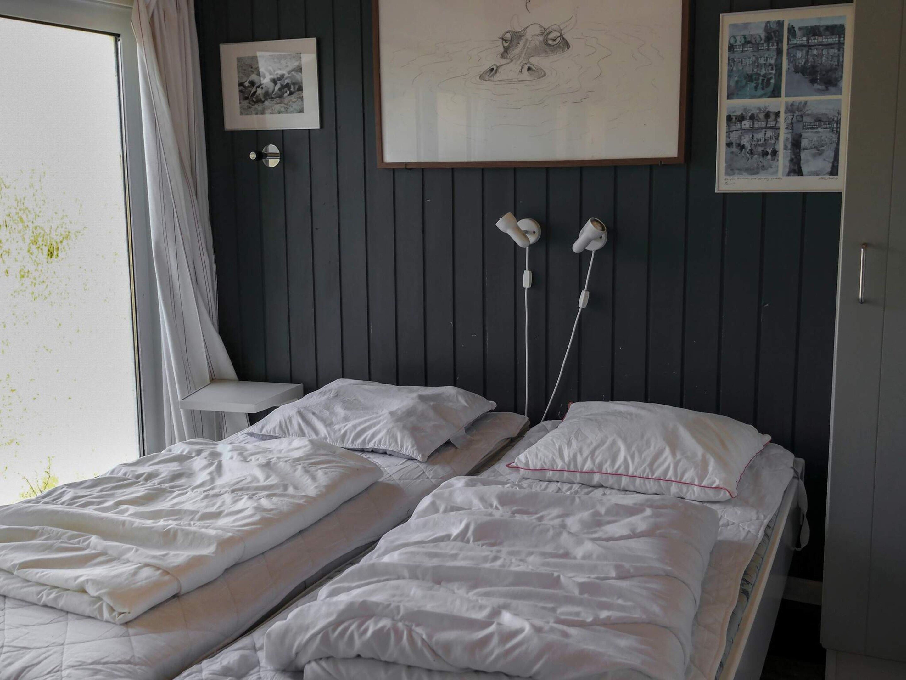 A room with two beds, a small table, and a door. There are wall-mounted lights and framed pictures.