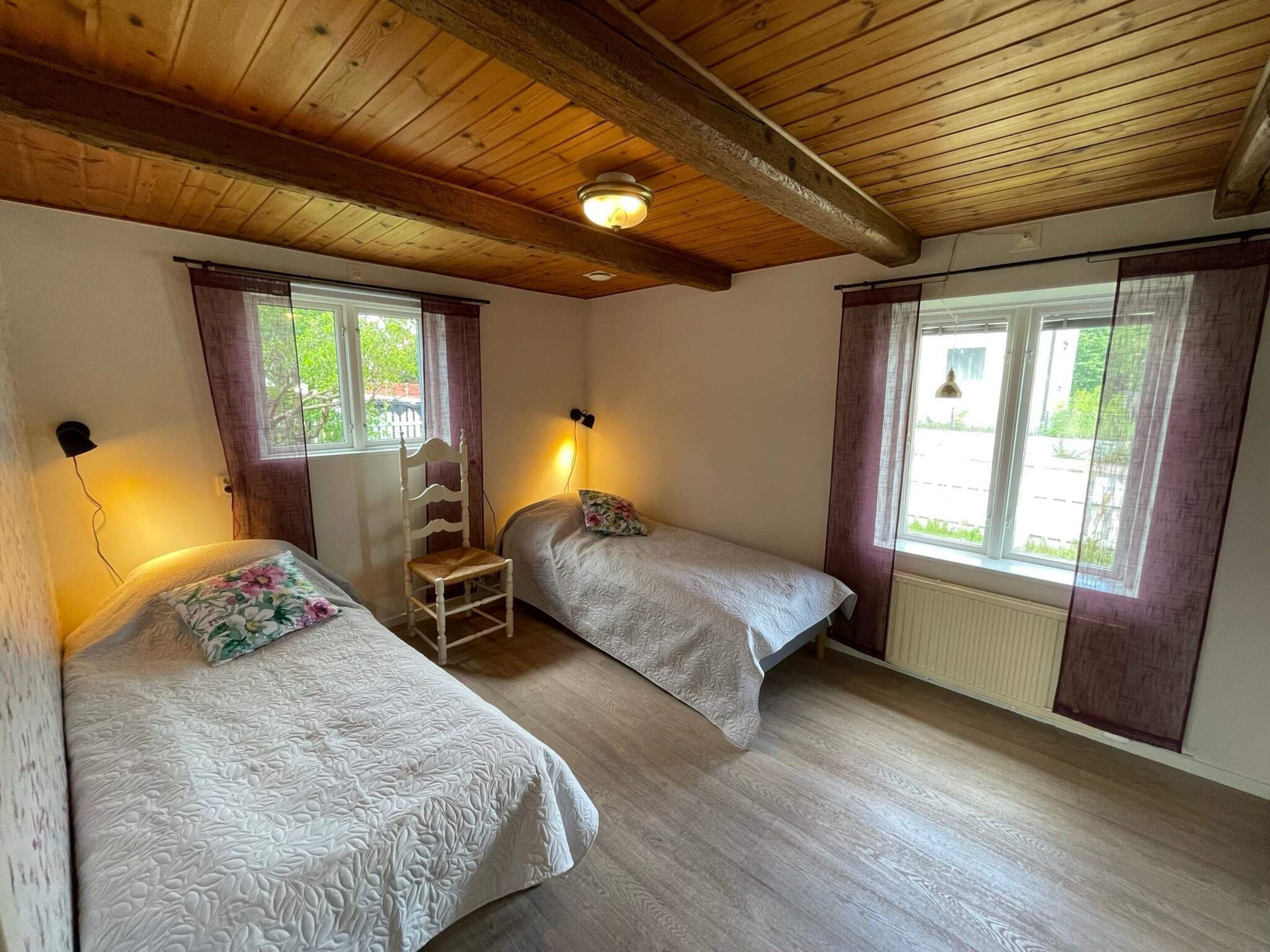 A room with two beds, a wooden ceiling, and windows with curtains.