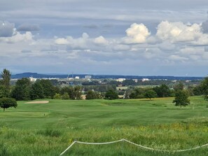 Un terrain de golf entouré d’une clôture en treillis métallique, bordé d’arbres, avec une vue lointaine sur la ville, le tout sous un ciel nuageux.