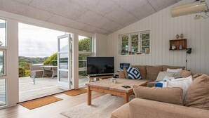 Ett rymligt vardagsrum med en stor TV, ett träbord och en beige soffa. Det finns en skjutdörr i glas som leder ut till en uteplats med ett bord och stolar.