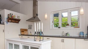 Ett kök med vita skåp, en köksö i mitten med glasluckor, ett fönster med utsikt över grönområde och en hängande lampa.