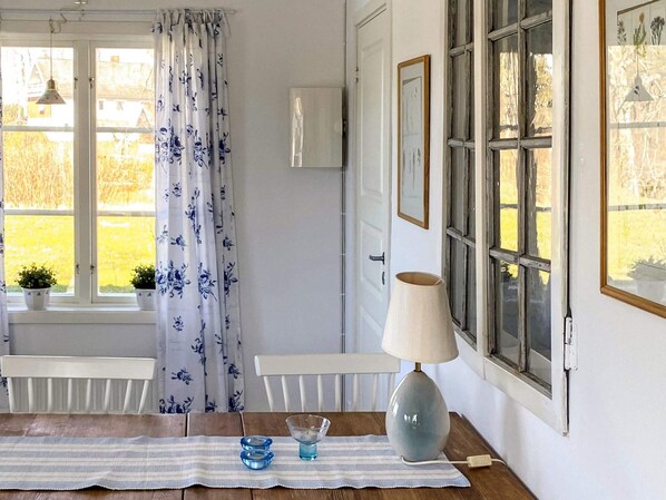 A room with a wooden table, a lamp, and a view of a garden through a window with blue floral curtains. - 4 Star Holiday Home in Bengtsfors (Bengtsfors)