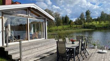 A wooden deck with a glass enclosure overlooking a lake, surrounded by greenery.