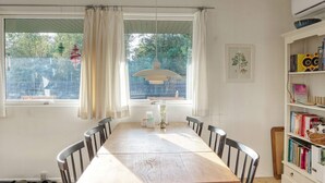 A dining area with a wooden table and chairs, a window with curtains, a pendant light, and a bookshelf with books and decorative items. - 4 Person Holiday Home in Vaeggerlose (Vaeggerlose)