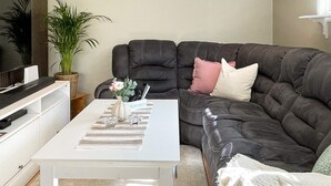 A living room with a dark grey sofa, a white coffee table, a TV on a stand, a potted plant, and a striped rug.