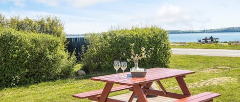 Una mesa de picnic de madera con bancos, un florero con flores y dos copas de vino en una terraza junto a un lago.