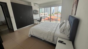 Un dormitorio con cama, escritorio, silla, armario y vistas a la ciudad a través de amplios ventanales.