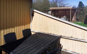 Eine Holzdauerdecke mit Tisch und Stühlen, ein überdachter Bereich und im Hintergrund Bäume sowie ein Gebäude.
