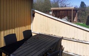 A wooden deck with a table and chairs, a covered area, and a view of trees and a building in the background. - 6 Person Holiday Home in Hojby (Hojby)