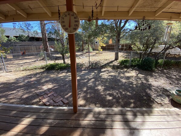 A covered outdoor area with a wooden bench, a hanging thermometer, and a fenced garden.