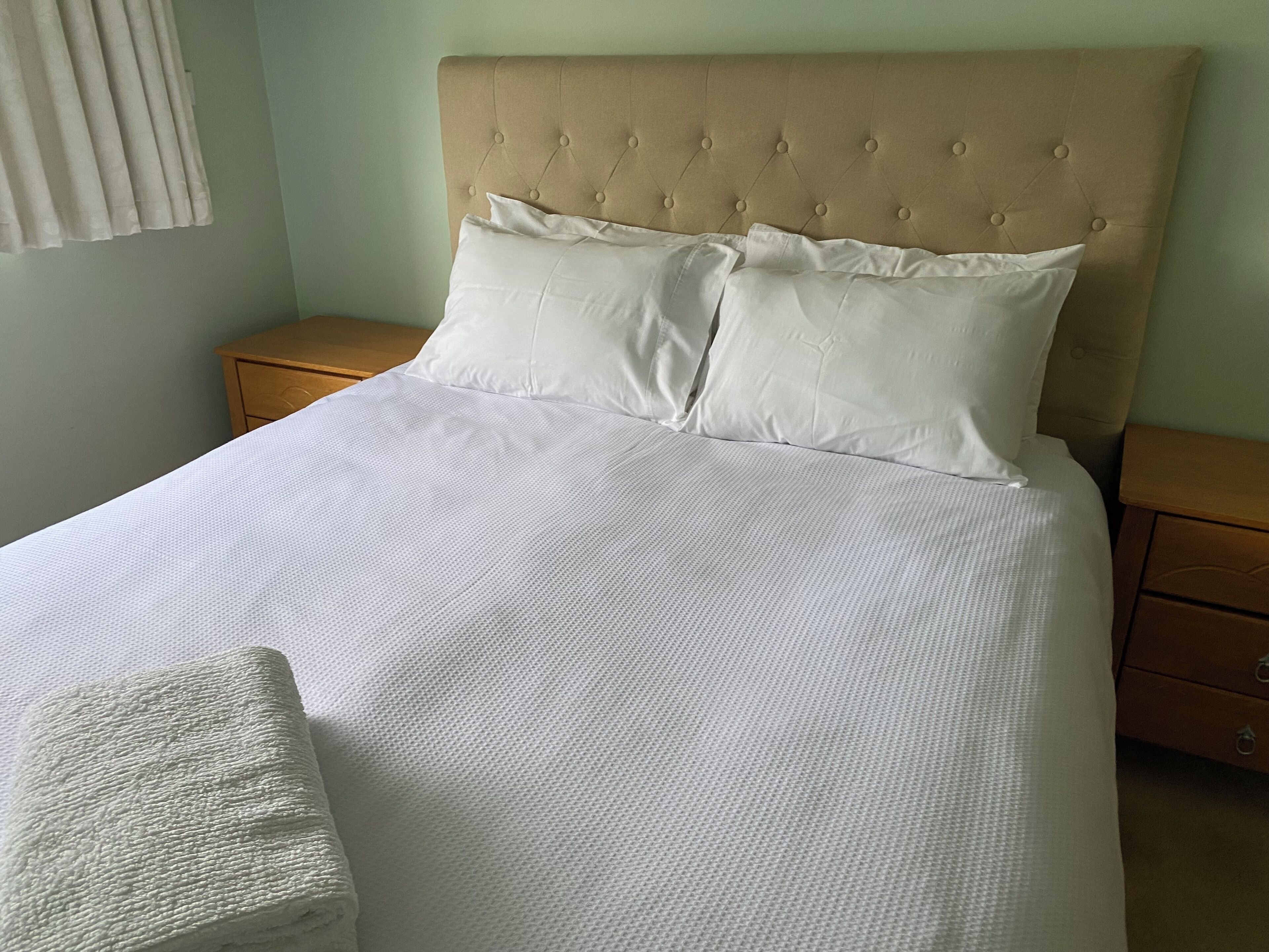 Una cama bien hecha con sábanas blancas, un cabecero capitonado y una mesita de noche de madera con un cajón.