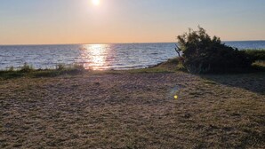 Ein Strand mit untergehender Sonne über dem Wasser, einer grünen Fläche und einigen Büschen.
