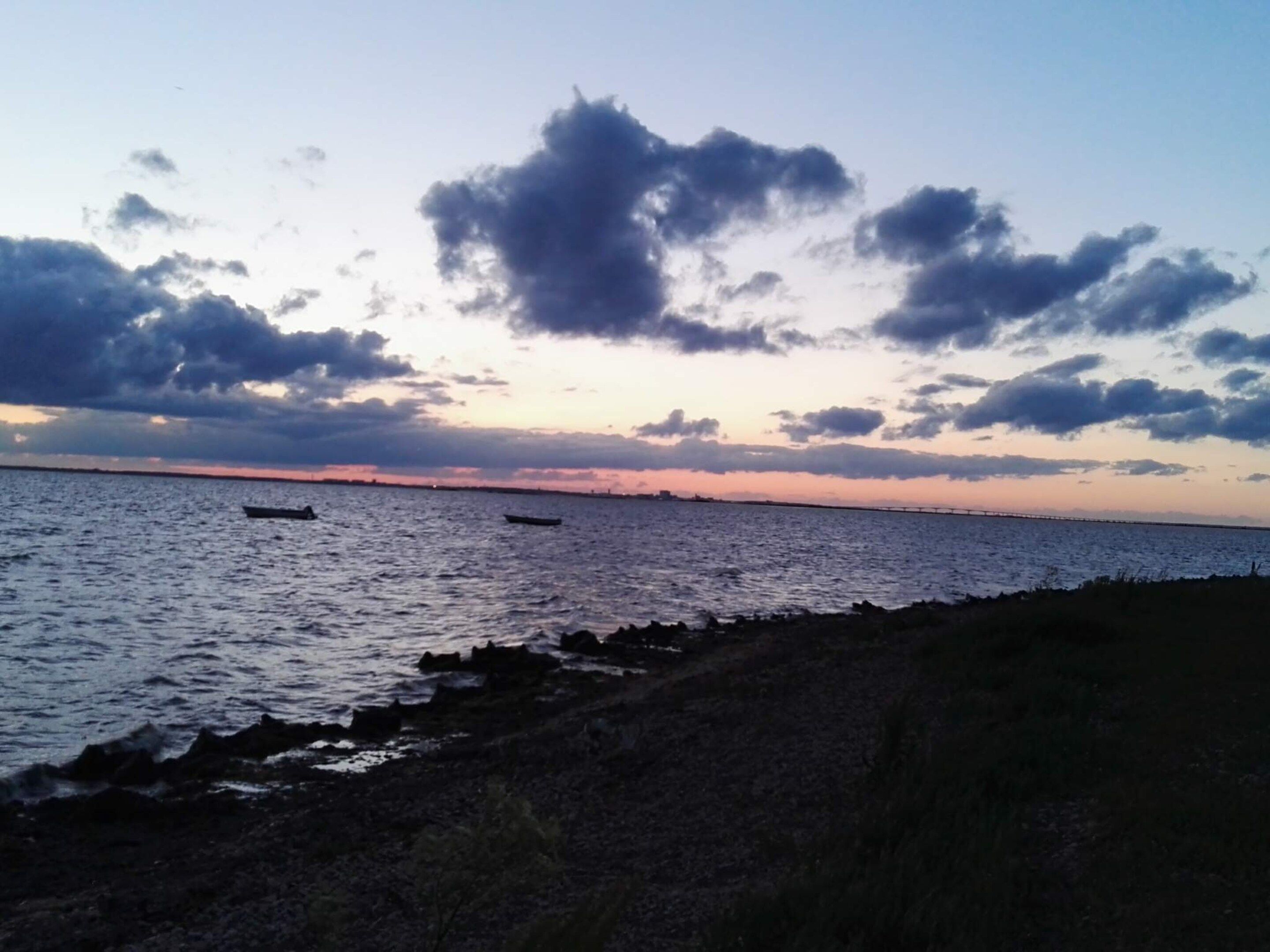 Paisaje costero con aguas tranquilas, costa rocosa y un puente distante.