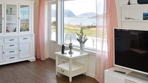 A room with a large window offering a view of a body of water and hills, a television on a stand, a cabinet with glass doors, and a small table with a lamp and a plant. - 8 Person Holiday Home in Leinoy-by Traum (Heroy)