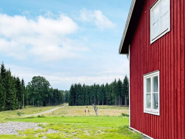 A red building with a white window, surrounded by a grassy field and a forest.