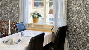En spiseplads med et bord dækket til fire personer, stole, et vindue med gardiner, en lampe og en vase med blomster.