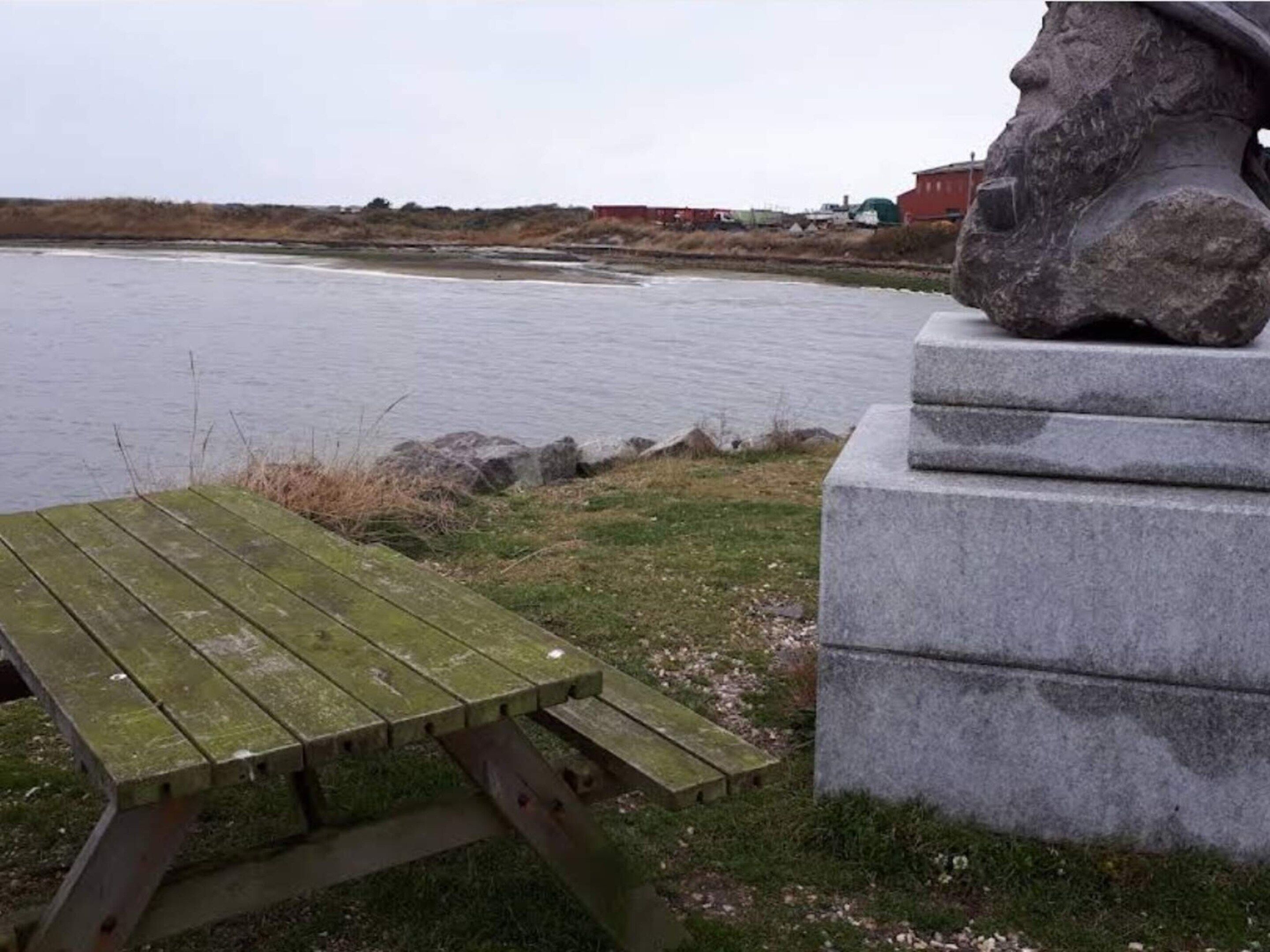A stone monument with a carved head, a wooden picnic table, and a body of water in the background.