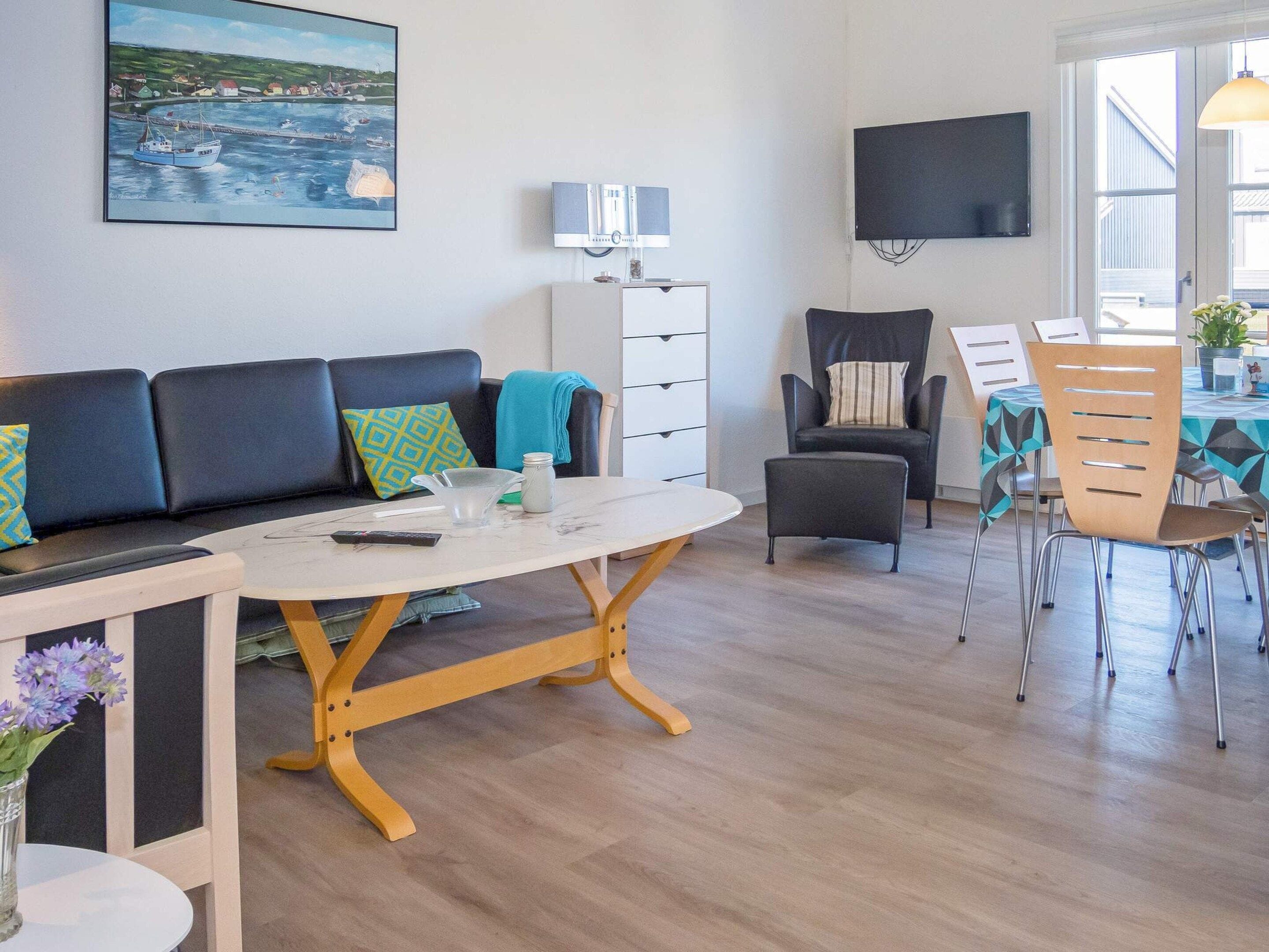 A modern living room with a sofa, a coffee table, a dining table with chairs, a TV mounted on the wall, and a painting of a coastal scene.