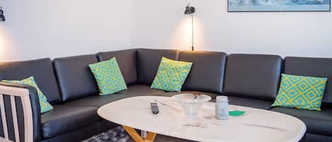 Un salon moderne avec un canapé d’angle, une table basse ronde et un tableau représentant un paysage côtier.