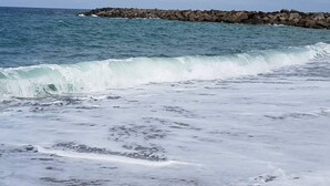 Uma praia com ondas quebrando na costa e um paredão rochoso ao fundo.