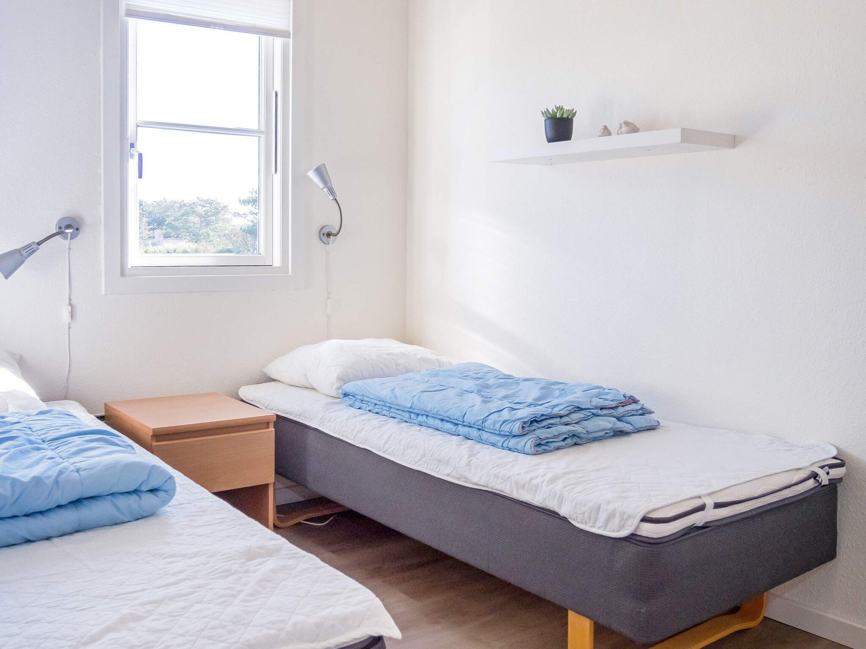A bedroom with two single beds, a bedside table, a lamp, a shelf with a plant, and a window.