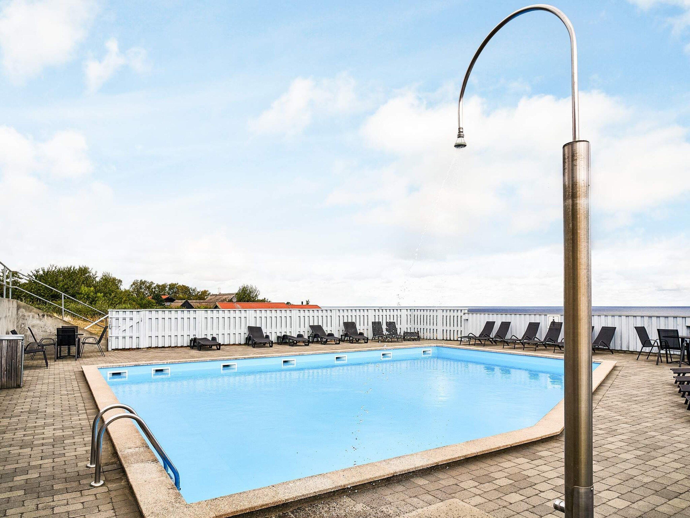 A rectangular swimming pool with a metal handrail on one side, surrounded by lounge chairs and a white fence.