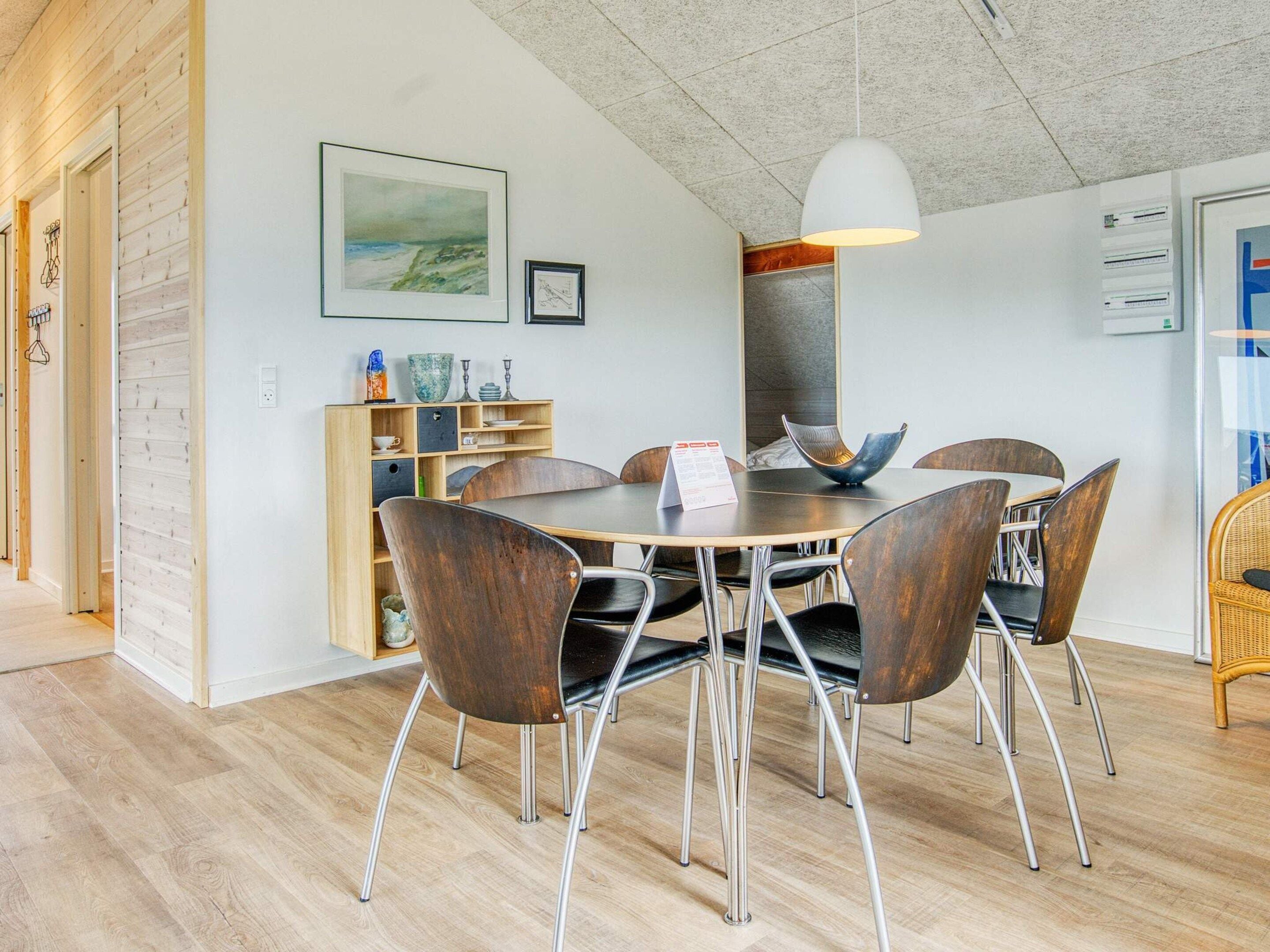 A dining area with a round table, six chairs, a white pendant light, and a wooden bookshelf with various items.