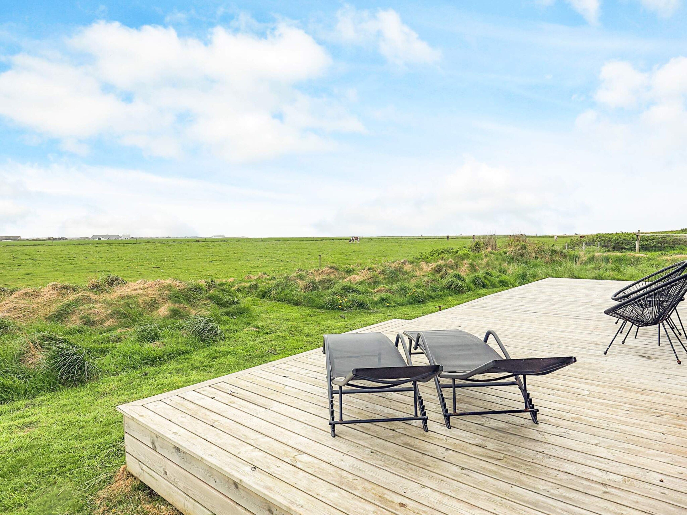 A wooden deck with two lounge chairs and a table, overlooking a grassy field with a clear sky.