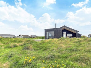 Une maison moderne à l’extérieur sombre, dotée de grandes fenêtres et d’une cheminée. Elle est entourée de dunes herbeuses et d’autres maisons qui se dessinent à l’arrière-plan.