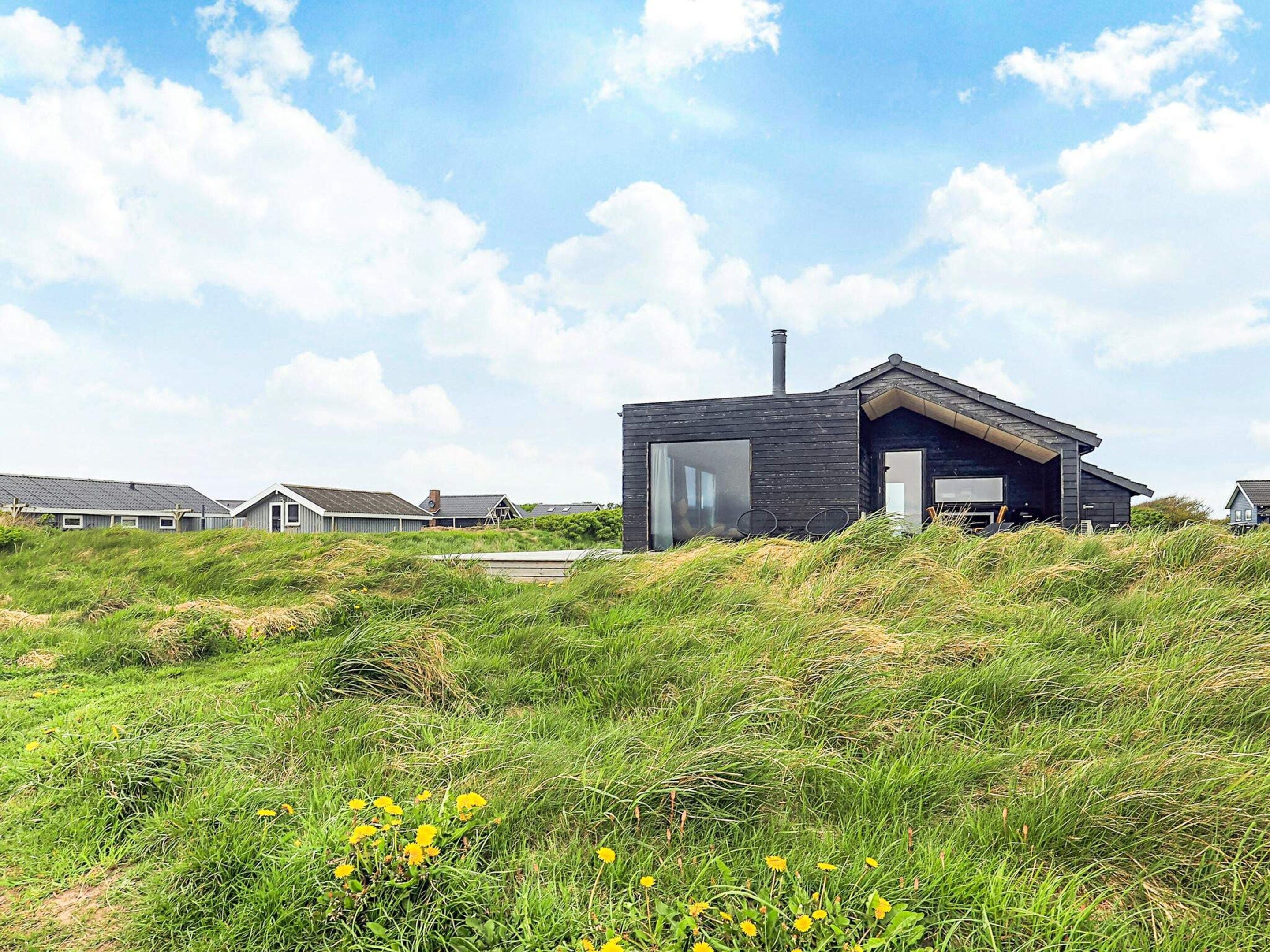A modern house with a flat roof, large windows, and a chimney, surrounded by grassy dunes and other houses.