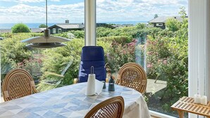 Un comedor con una mesa dispuesta para cuatro personas, rodeado de sillas de mimbre, con vistas a un jardín exuberante y una vista panorámica de casas y el mar.