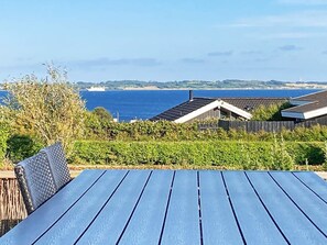 A view of a coastal area with a wooden table in the foreground, a fenced garden, and a distant view of the sea and a ship. - 8 Person Holiday Home in Ebberup (Ebberup)