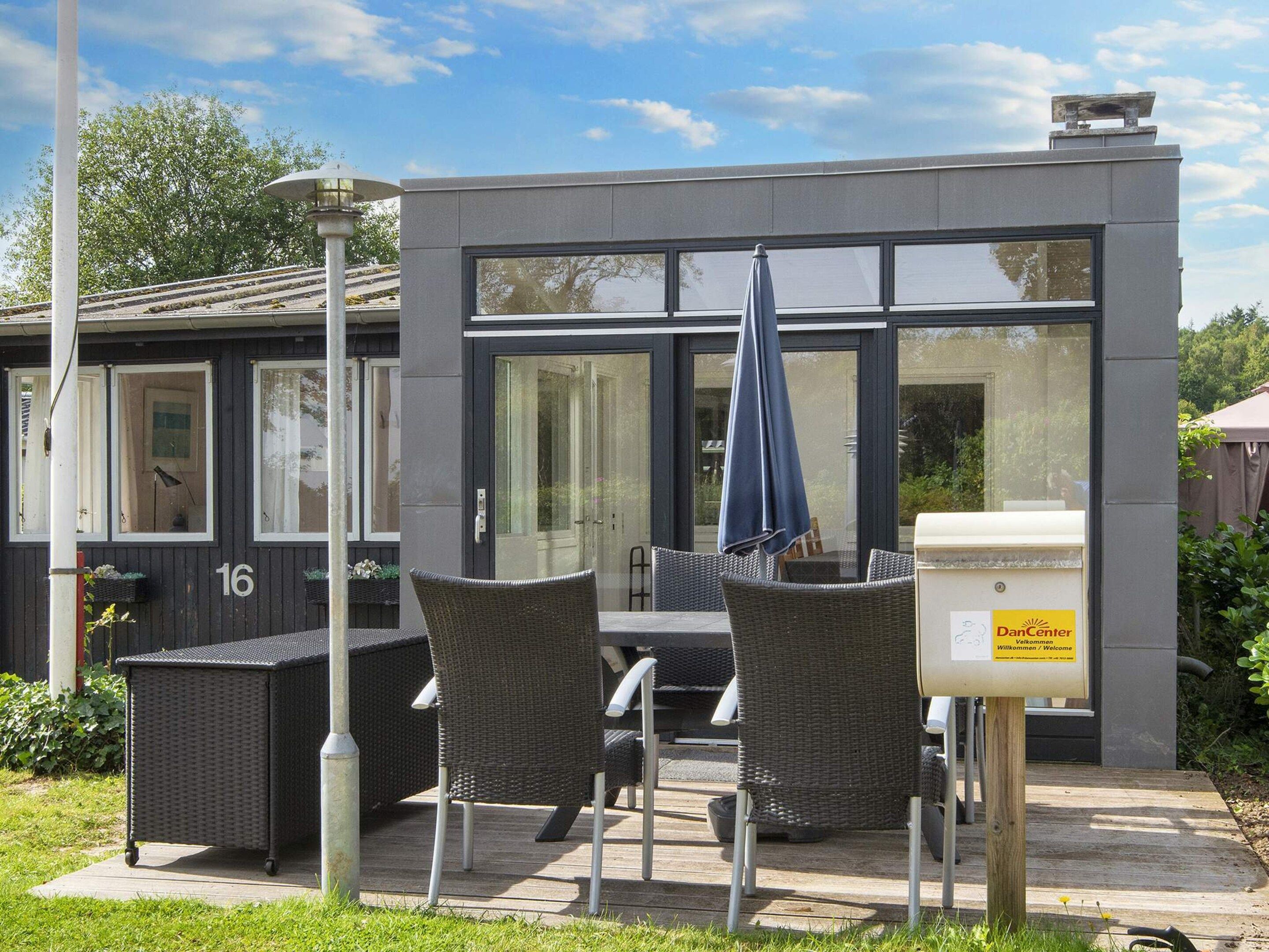 A modern building with a large glass window, outdoor seating with wicker chairs and a table, and a sign indicating a welcome area.