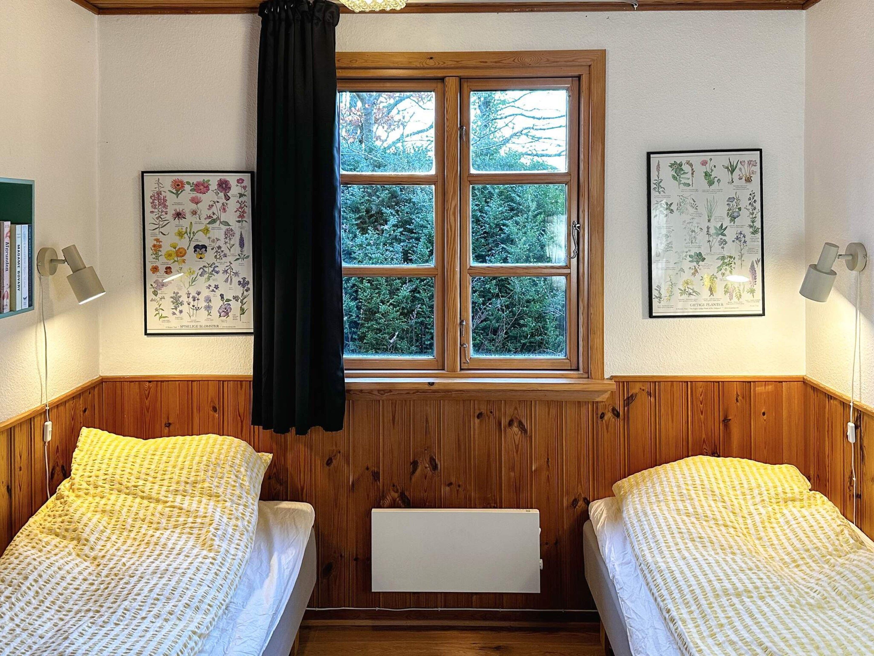 Ein Zimmer mit zwei Betten, Holzvertäfelung, einem Fenster mit Blick auf Bäume und zwei gerahmten Pflanzenabbildungen an den Wänden.