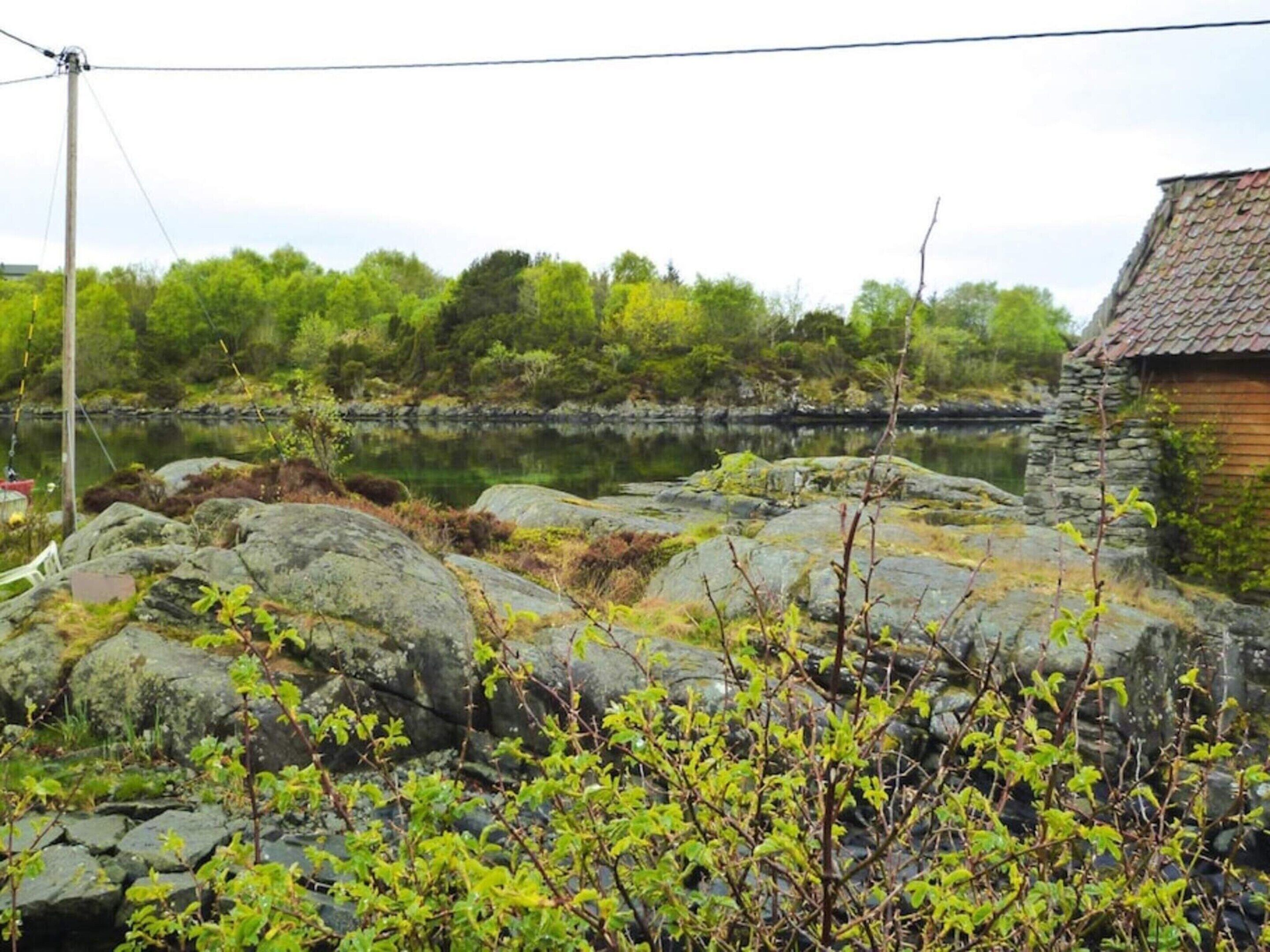 A rustic stone building with a tiled roof, situated next to a calm body of water, surrounded by lush greenery and large rocks.