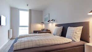 A modern bedroom with a large bed, a bedside table, a nightstand, a window with curtains, and a wall-mounted light fixture.