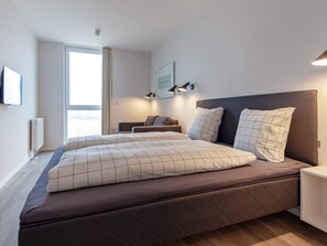 A modern bedroom with a large bed, a bedside table, a nightstand, a window with curtains, and a wall-mounted light fixture.