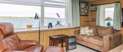 A cozy room with a leather chair, a brown sofa, and a small table with a lamp. A window offers a view of the water.