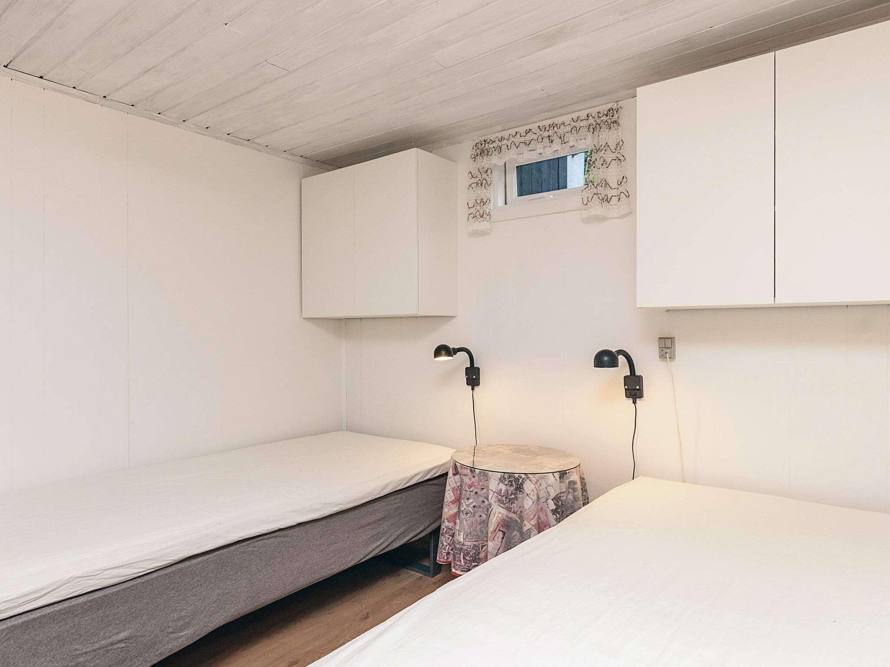 A small, minimalist room with two single beds, a small round table, and a window with a decorative frame.