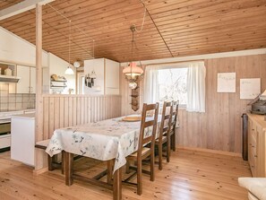 Interior em madeira com uma mesa de jantar posta para quatro, área de cozinha e janela com cortinas.
