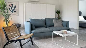 A modern living room with a grey sofa, a woven chair, a white coffee table, and a grey rug. There is a white wardrobe, a plant, and a framed artwork on the wall.