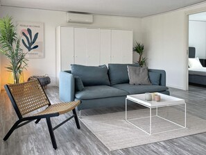 A modern living room with a grey sofa, a woven chair, a white coffee table, and a grey rug. There is a white wardrobe, a plant, and a framed artwork on the wall. - 4 Star Holiday Home in Borgholm (Borgholm)