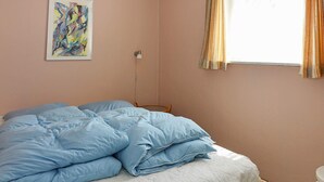 Een slaapkamer met een bed, nachtkastje, lamp, raam met gordijnen en een schilderij aan de muur.