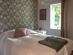 Un dormitorio con papel tapiz floral, una cama con cubrecama acolchado, una mesita de noche con lámpara y una ventana con cortinas.